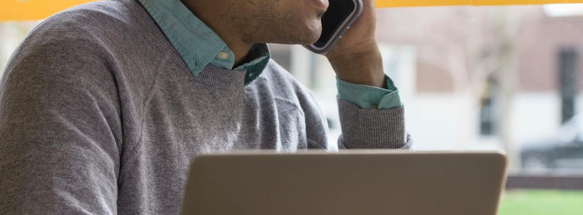 Smiling student talking on a cellphone while using a laptop at a café.