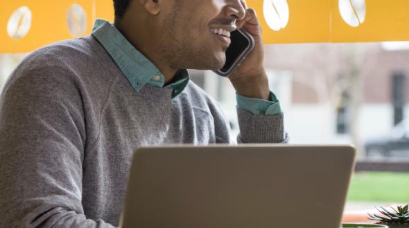 Smiling student talking on a cellphone while using a laptop at a café.