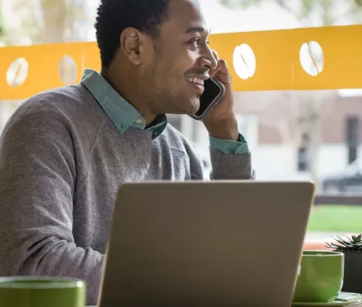 Smiling student talking on a cellphone while using a laptop at a café.
