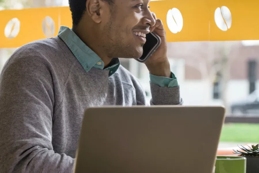 Smiling student talking on a cellphone while using a laptop at a café.